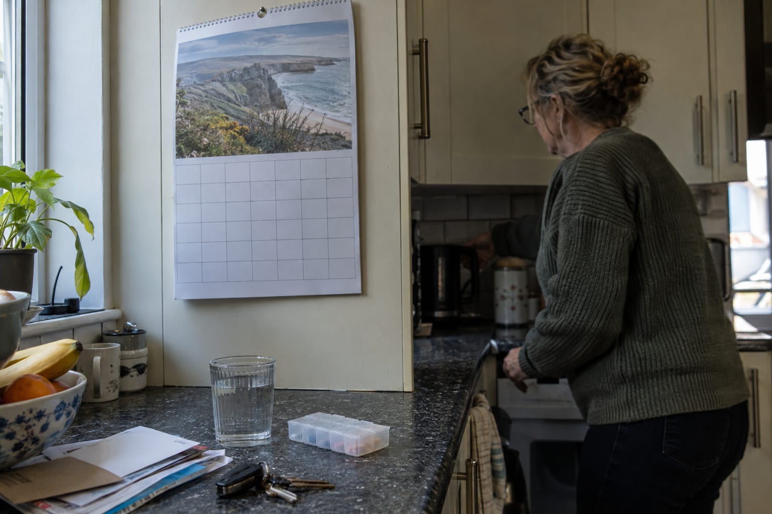 A wall calendar on a kitchen cupboard above a weekly pill organiser and a glass of water, with a woman at the kettle in the background