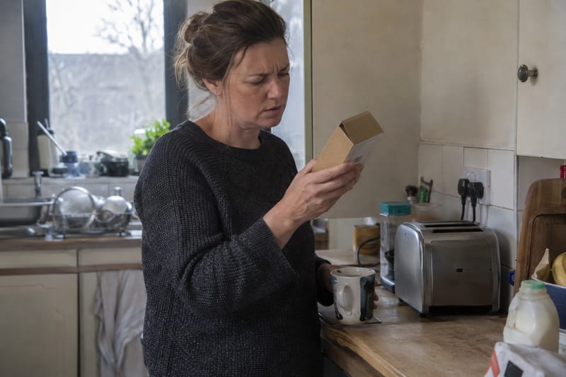 A woman in a British kitchen frowning at the back of a small cardboard box while holding a mug