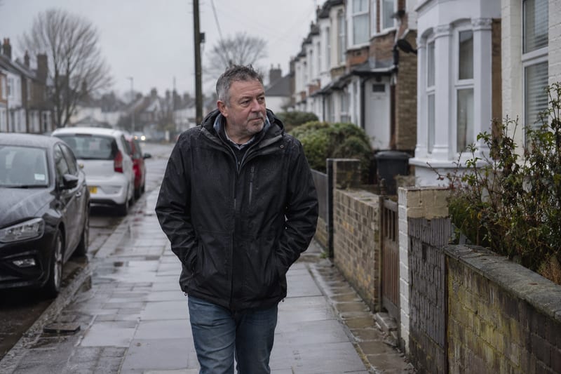 A man in a dark waterproof jacket walking along a wet terraced street on a grey morning